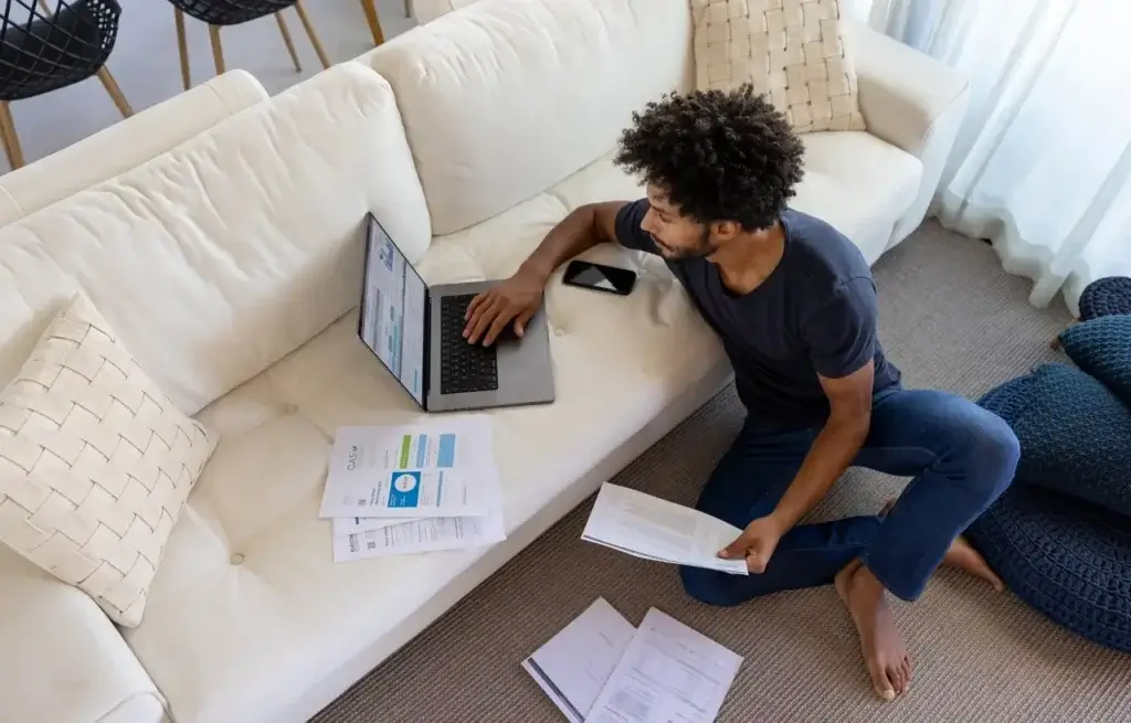 homem com contas na mão sentado no chão mexendo no computador que está sobre o sofá para representar "como entender a conta de luz"