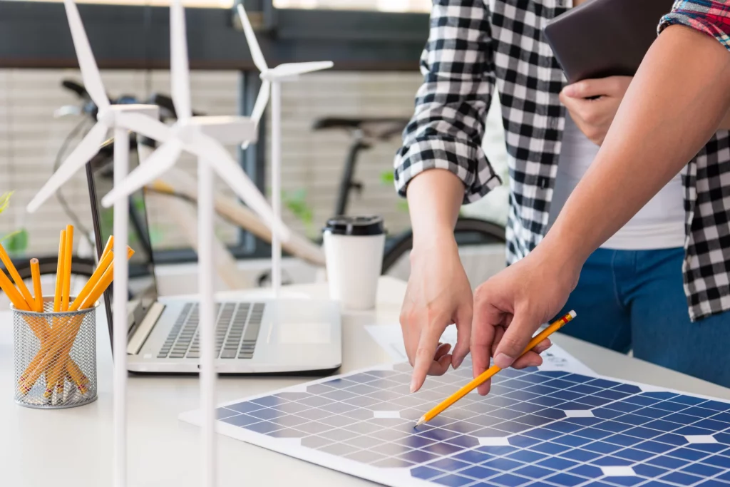 Duas pessoas apontam para uma placa de energia solar em uma mesa. Há um laptop, vários lápis amarelos em um escritório